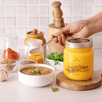 A2 ghee jar with a spoonful of content over a bowl of soup, surrounded by dal, spices and herbs.