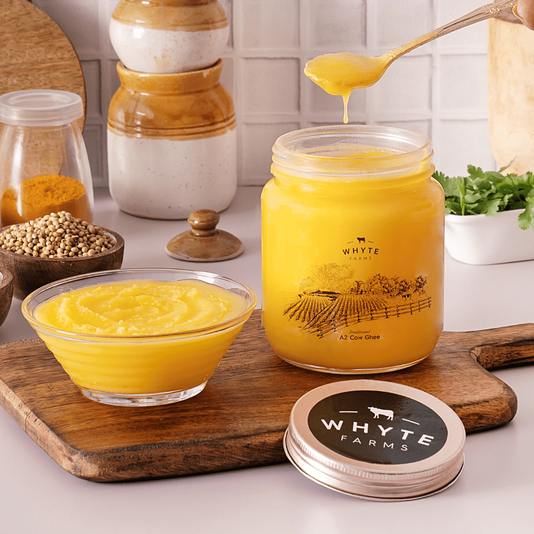 Jar of rich, grainy ghee with a spoon, bowl, and wooden cutting board on a white surface.