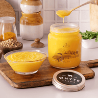 Jar of rich, grainy ghee with a spoon, bowl, and wooden cutting board on a white surface.