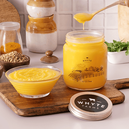 Jar of rich, grainy ghee with a spoon, bowl, and wooden cutting board on a white surface.